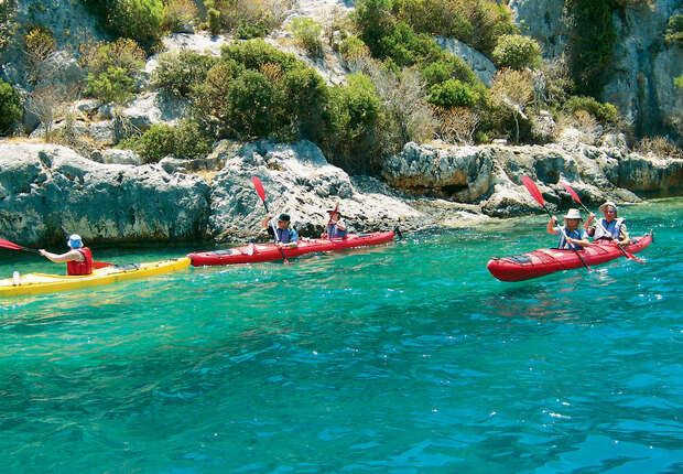 Kayaks with paddlers move through clear turquoise water near rocky cliffs adorned with green shrubs, providing a vibrant, adventurous setting.